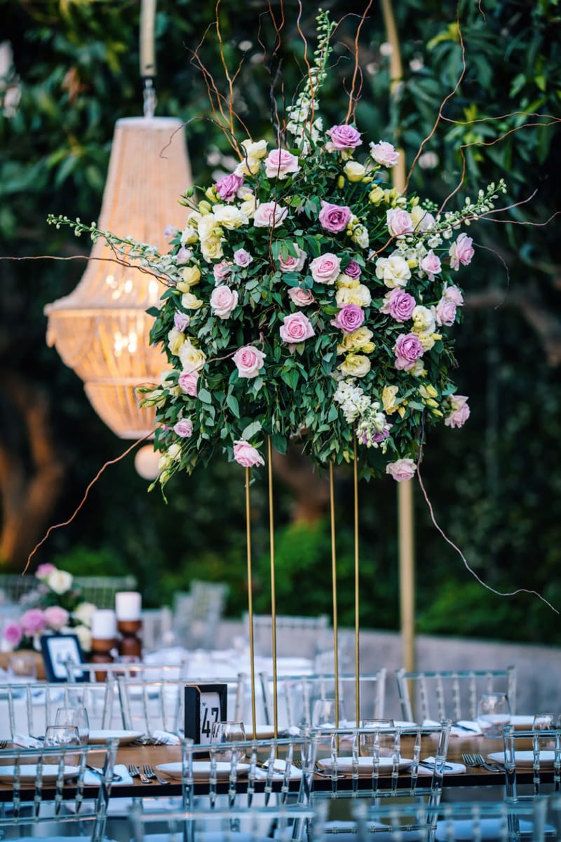 Tall arrangement of pink and white roses on a gold stand beside a beaded chandelier, over set tables with clear chairs