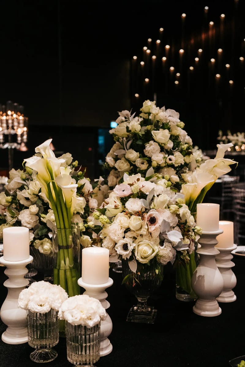 White roses, calla lilies and anemones beside white pillar candles and glass vases on a dark background