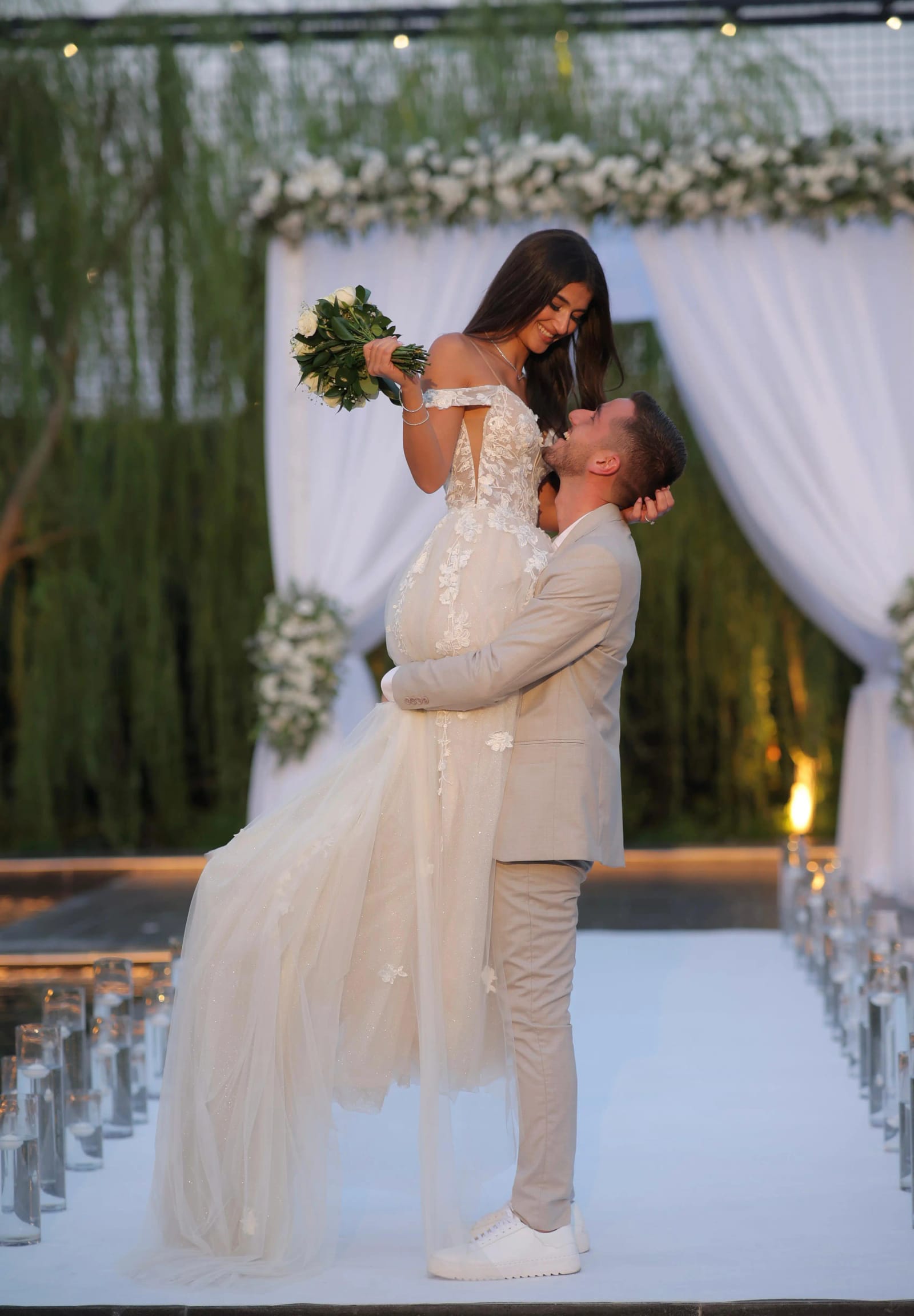 A groom lifting the bride in front of a white draped chuppah with a flower garland and a candle-lined aisle