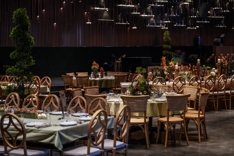 Hall with round tables in sage-green cloths, wooden chairs, a spiral topiary and a dark slatted wall with hanging lights