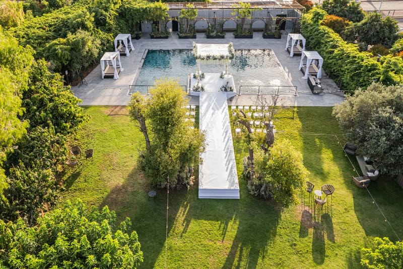 Aerial view of a lawn with a T-shaped white aisle leading to a pool with a white platform, among trees