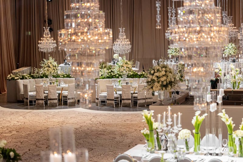 Lustres de cristal étagés au-dessus d'une table centrale dressée, callas et bougies au premier plan, rideaux beiges au fond