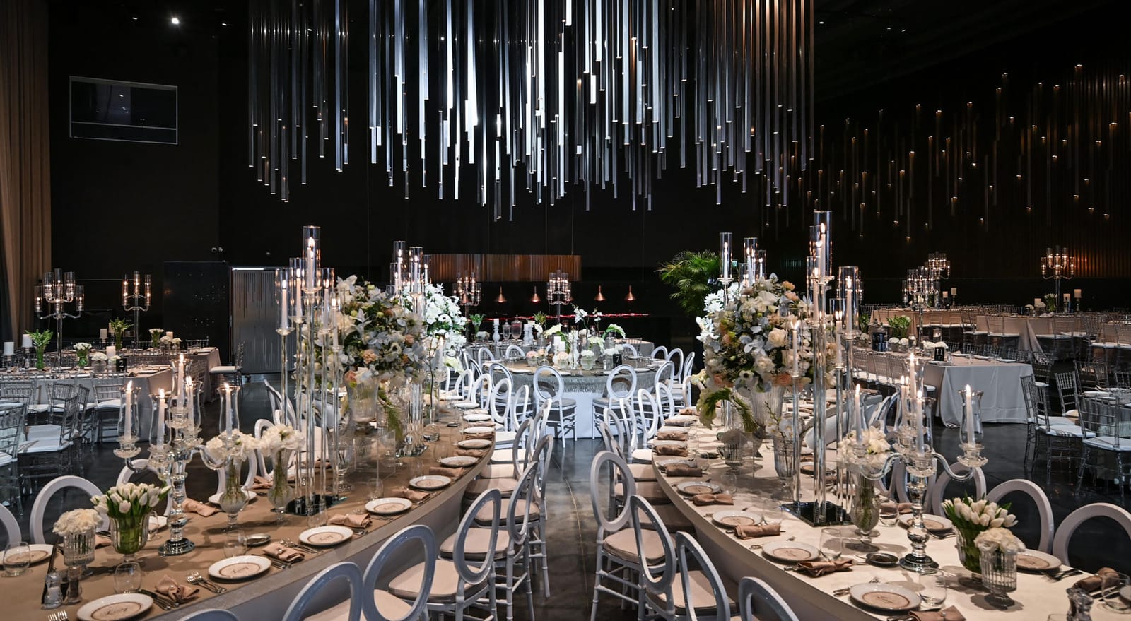Salle sombre avec longues tables, candélabres et fleurs blanches sous un plafond de tubes lumineux verticaux suspendus