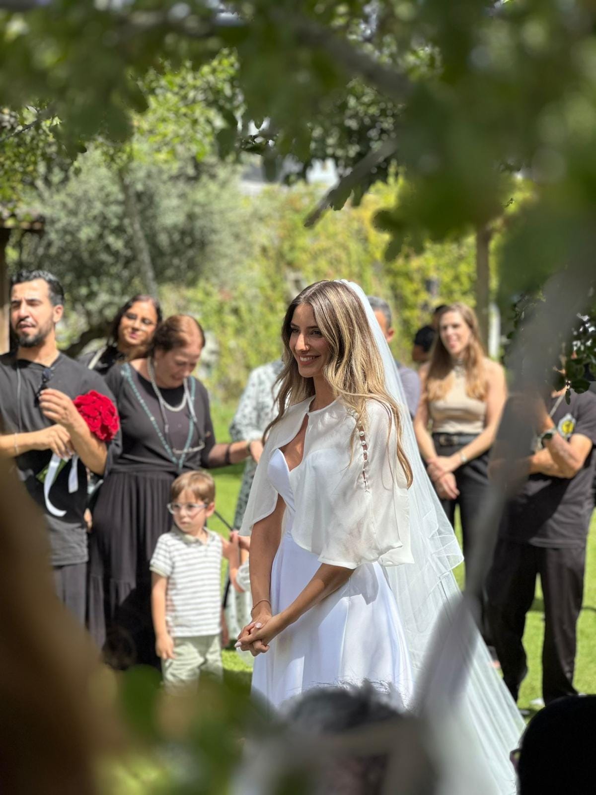 A smiling bride in a veil and white cape on the lawn, surrounded by guests and a small boy under trees
