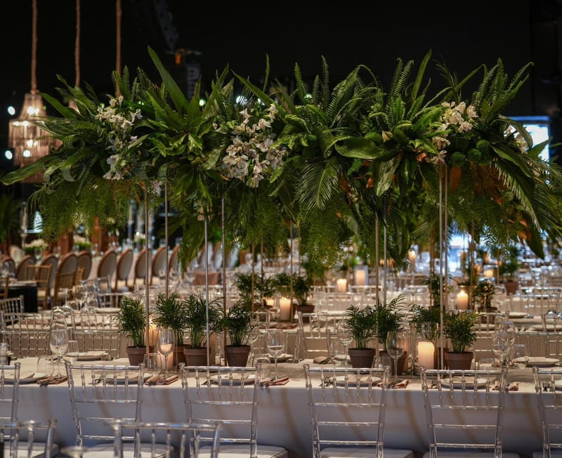 Hanging installation of ferns and white flowers above tables with potted palms, candles and clear chairs
