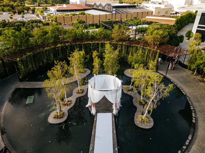 Vue aérienne d'un dais blanc drapé au bout d'une passerelle blanche sur une eau sombre, entre des îlots plantés d'arbres