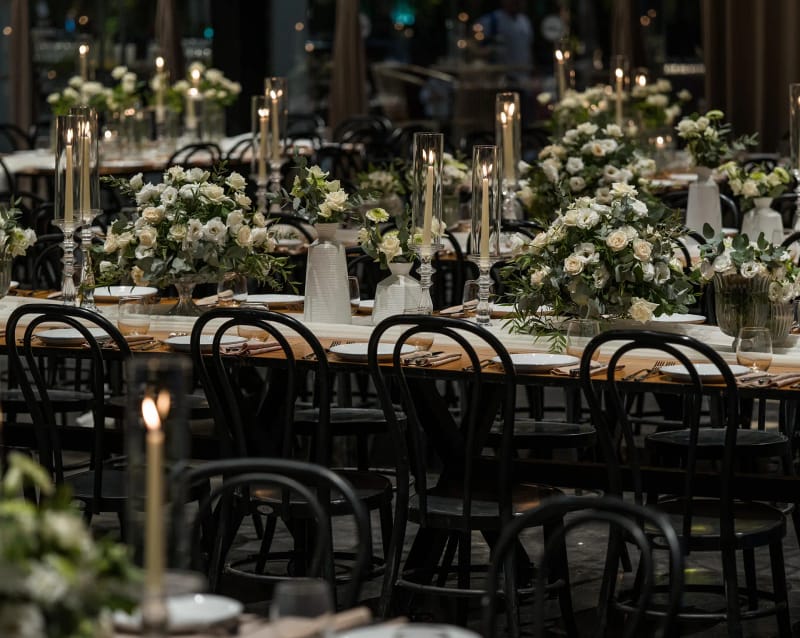 A row of green-and-white floral arrangements and tall candles along a long table with black chairs