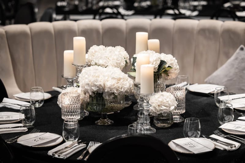 Gros plan d'hortensias blancs et de bougies pilier dans des vases en cristal sur une table noire, près d'une banquette capitonnée