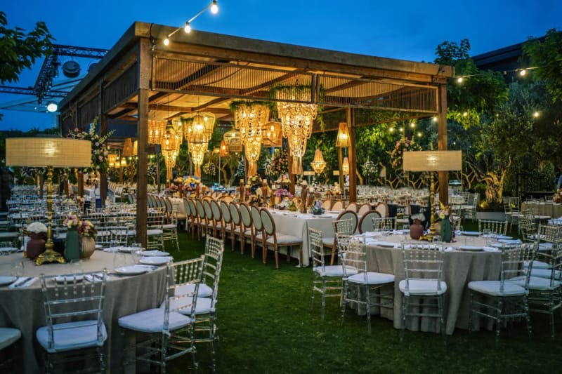 Wooden pergola with hanging gold beaded chandeliers over a long table and round tables on a lawn at blue hour