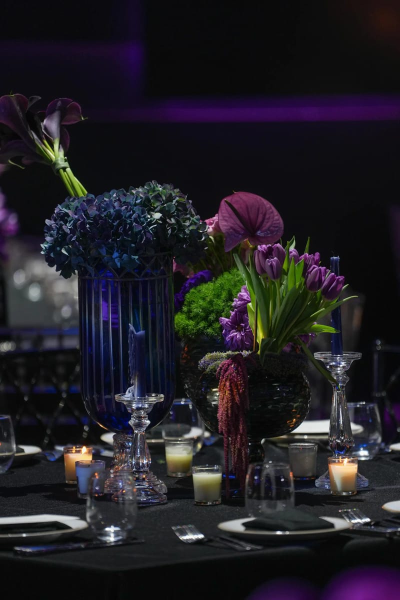Purple tulips, anthurium and hydrangea in a blue vase on a dark table with candles and place settings