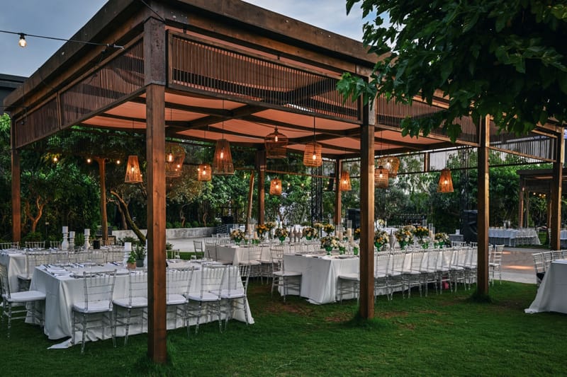 Wooden pergola with hanging lanterns above set tables on the lawn, among trees and string lights