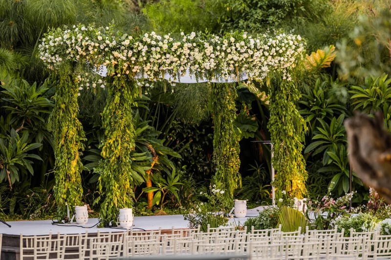 Round canopy covered in white flowers on greenery-wrapped columns, with white chairs and a lush garden