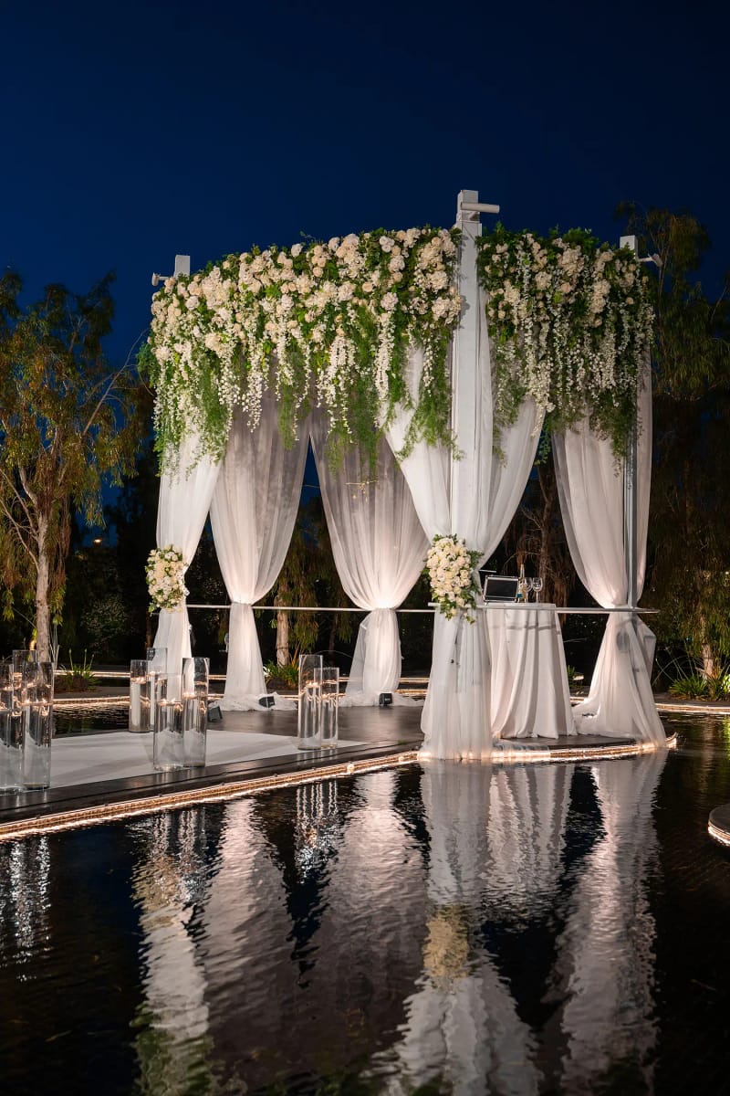 Houppa aux voiles blancs, fleurs blanches et feuillage en cascade au-dessus d’un bassin miroir, de nuit
