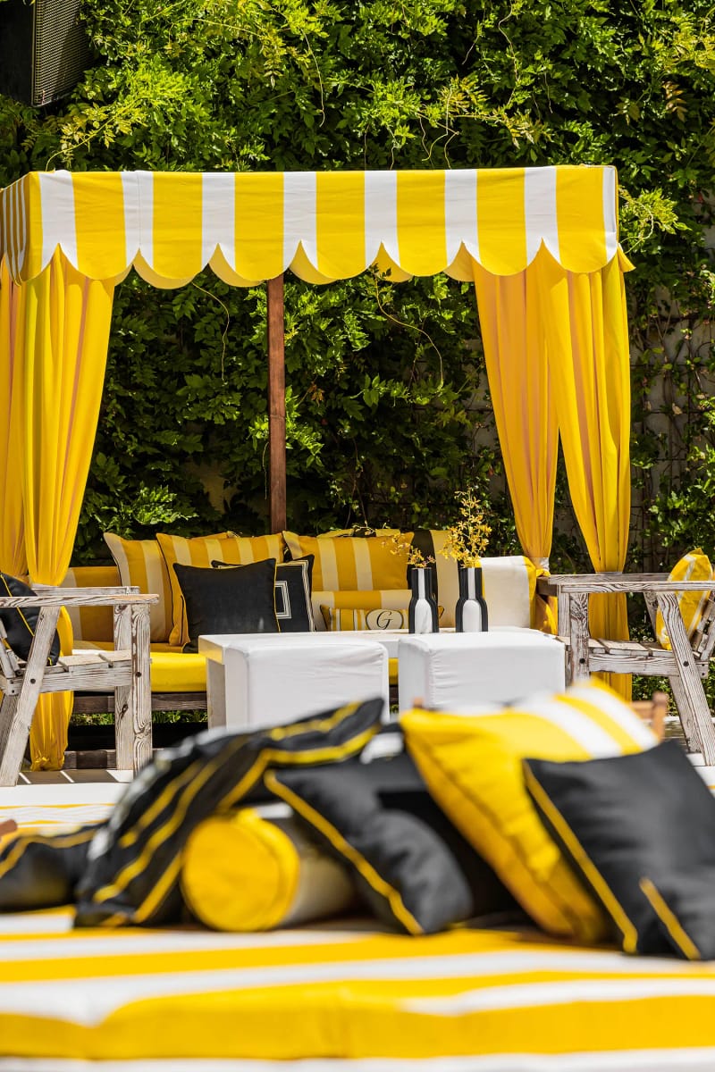 Yellow and white striped canopy with yellow curtains and a lounge area with black and yellow cushions, among greenery