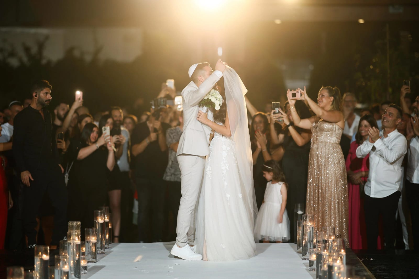 A couple kissing on a white platform in warm light while guests film from both sides of a candle-lined aisle