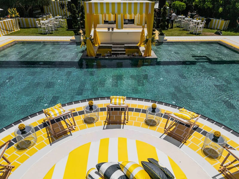 Curved yellow and white striped platform at the pool edge with deck chairs and tables, and a canopy across the water