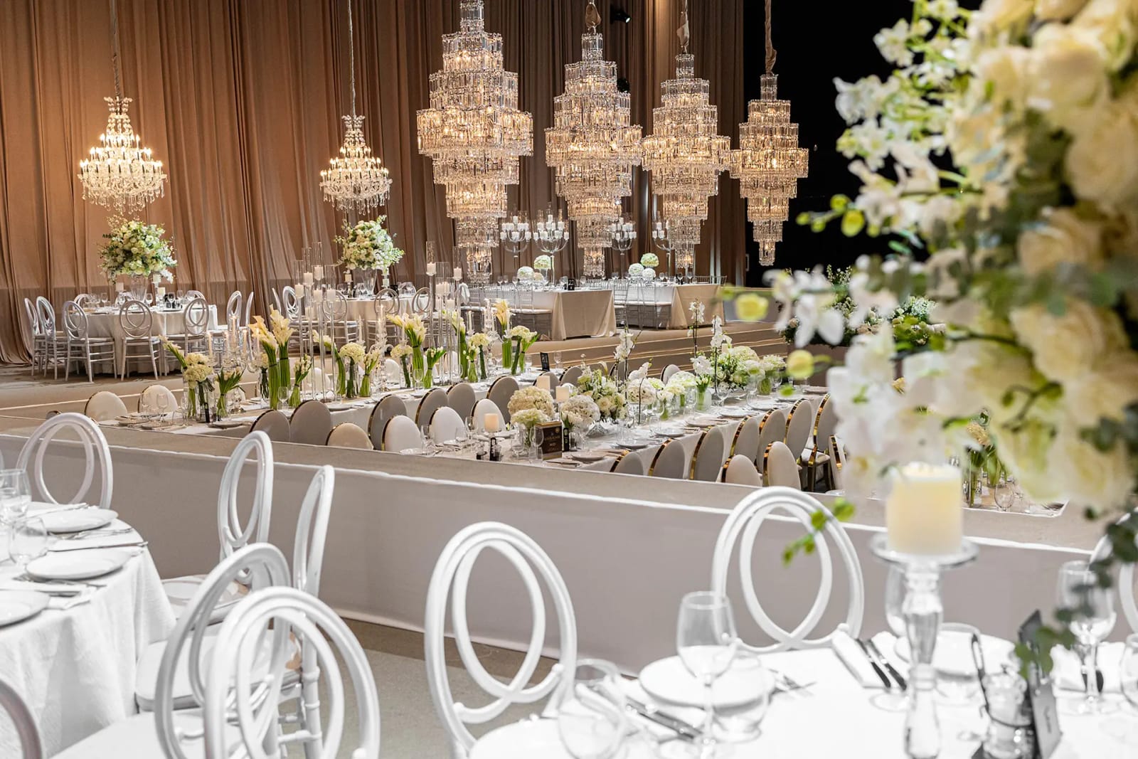 Hangar with tiered crystal chandeliers, beige drapes and a long white table with white chairs, in the Enchanted Ball design