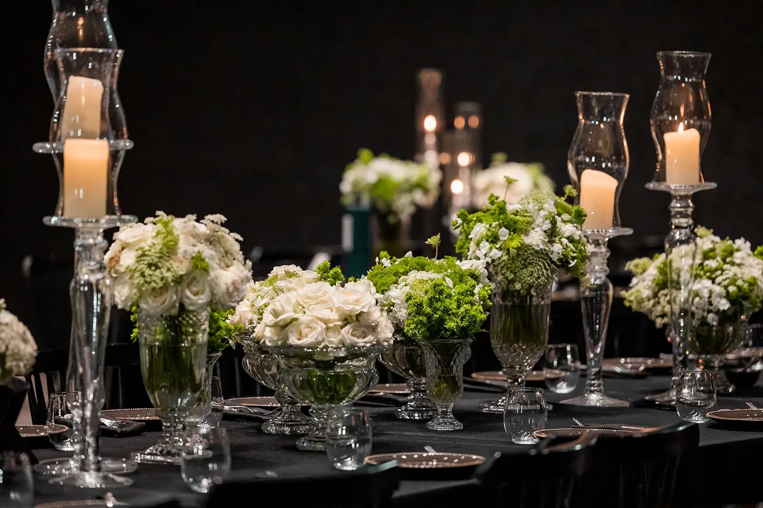 Rangée de coupes en verre garnies de fleurs blanches et vertes entre de hauts photophores sur une table sombre