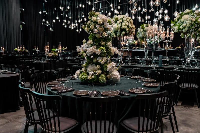 Table ronde nappée de vert foncé avec tour de fleurs au centre, chaises noires et ampoules en verre suspendues