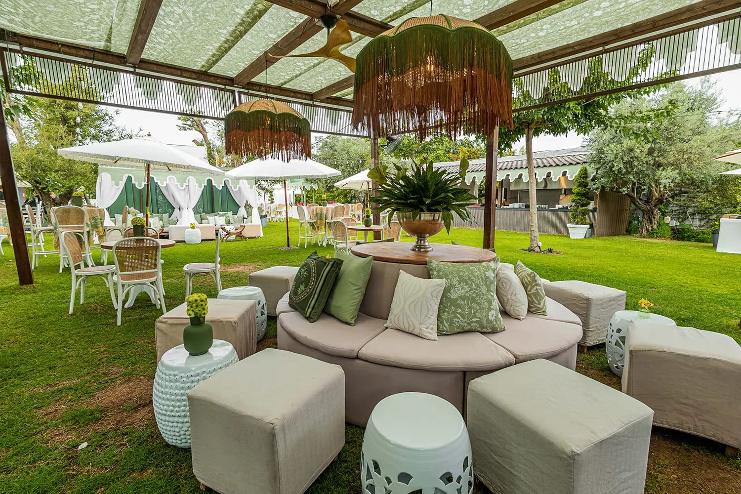 Salon sur pelouse avec canapé courbe crème, coussins verts, poufs et tabourets en céramique sous une pergola