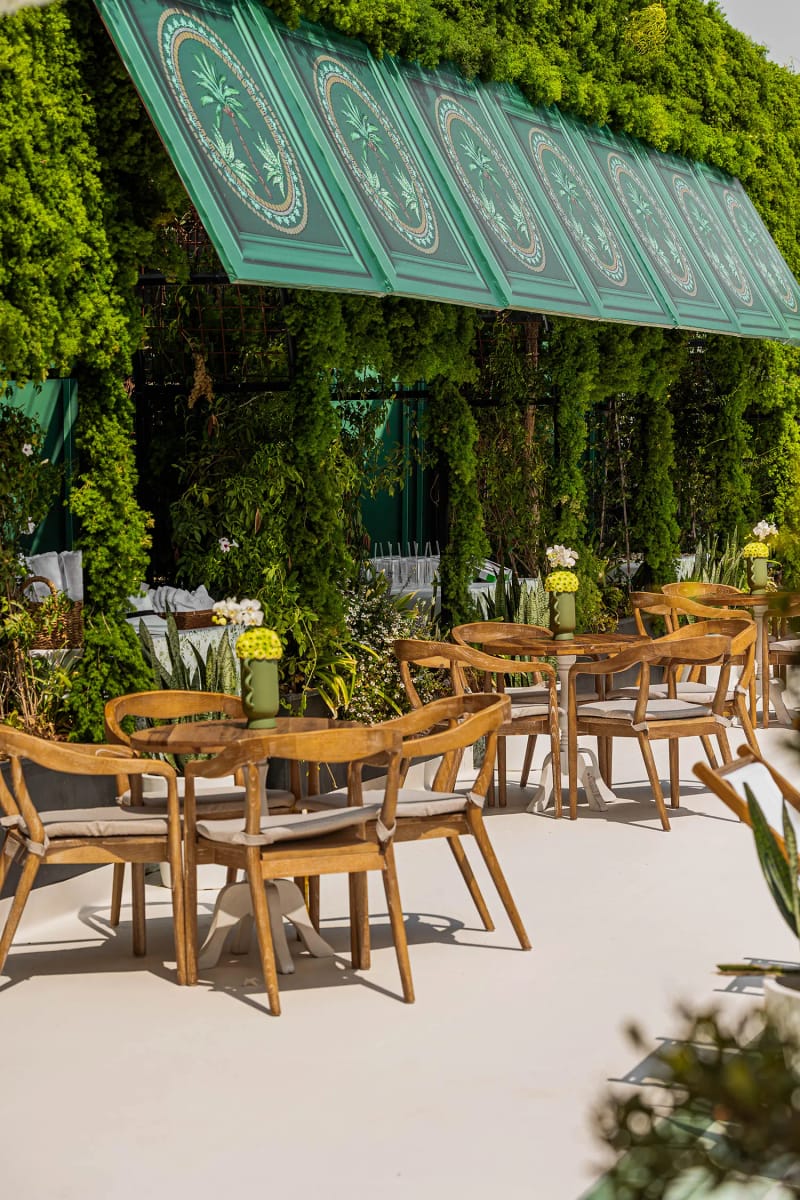 Tables et chaises en bois sur une terrasse claire sous un auvent vert à panneaux et un mur de lierre