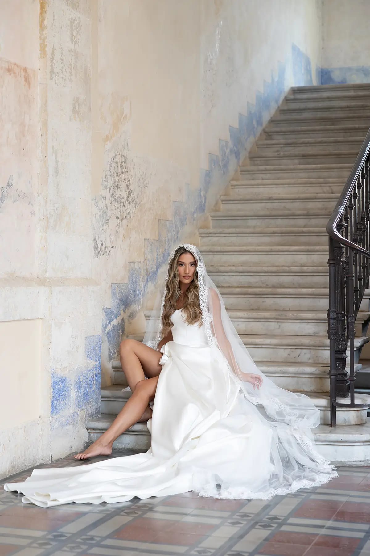 Une mariée pieds nus assise sur des marches en pierre près d'un mur de carreaux bleus, voile et robe étalés sur le sol