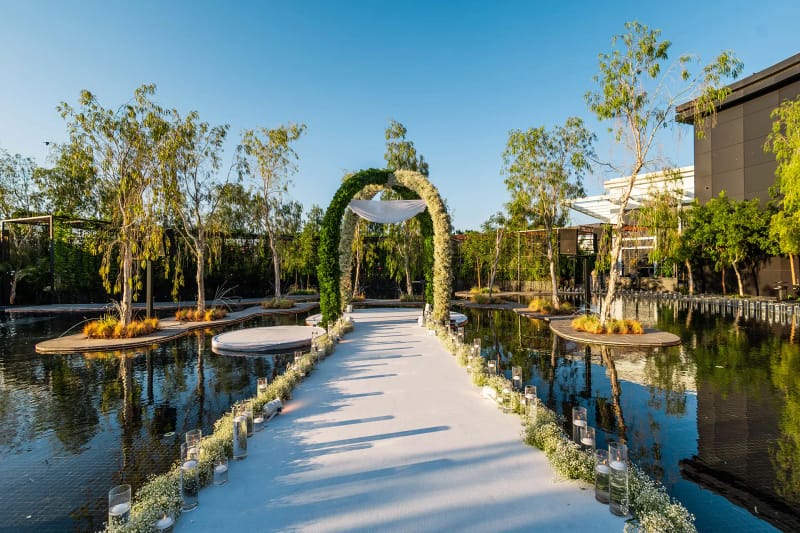 White aisle lined with flowers leading to a green arch, between islands with trees in the pond, on a clear day