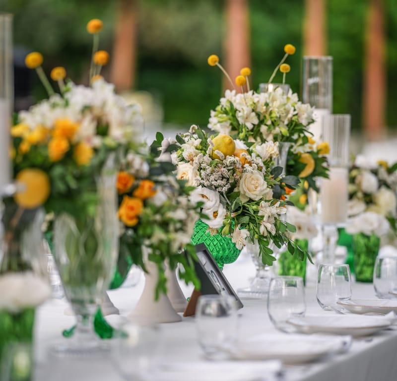 Bouquets of white and yellow flowers with foliage in glass vases, with place settings in the foreground