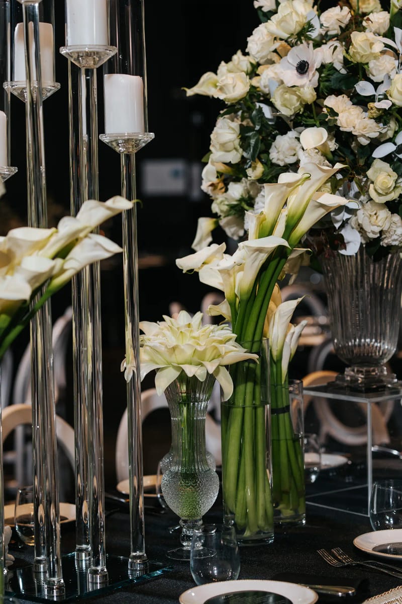 Close-up of white calla lilies in glass vases beside a white floral arrangement and tall glass candle holders