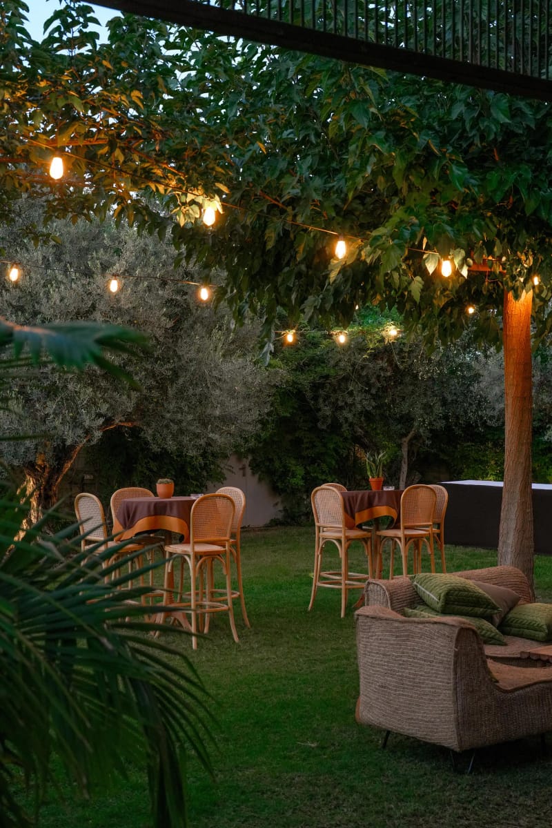 Garden corner at dusk with string lights, woven bar chairs and a high table beneath olive trees
