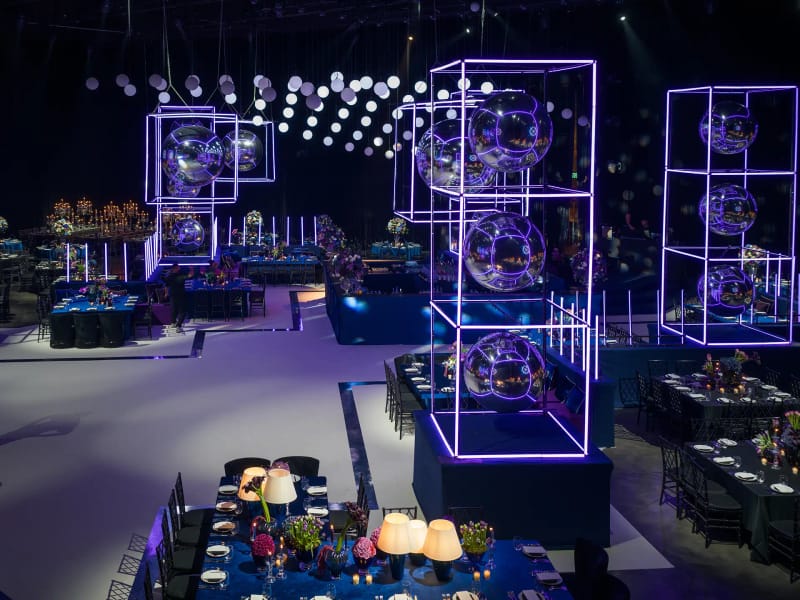 Dark hall with neon-lit cubes holding mirror spheres, hanging dots of light and blue-clothed tables around a white floor