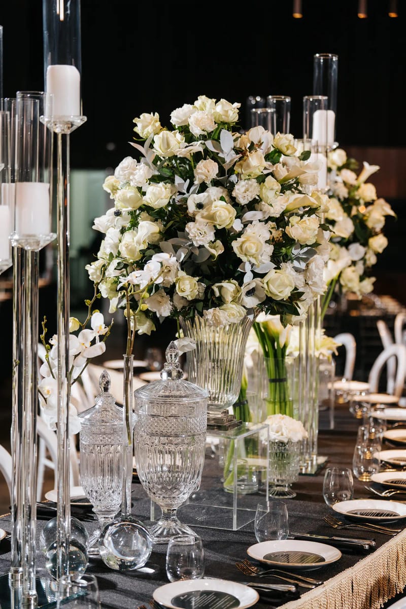 White rose bouquet on a tall glass stand with candles, crystal glasses and black plates on a table