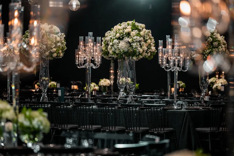 Candélabres et compositions de fleurs blanches sur de hauts pieds en verre au-dessus de tables sombres