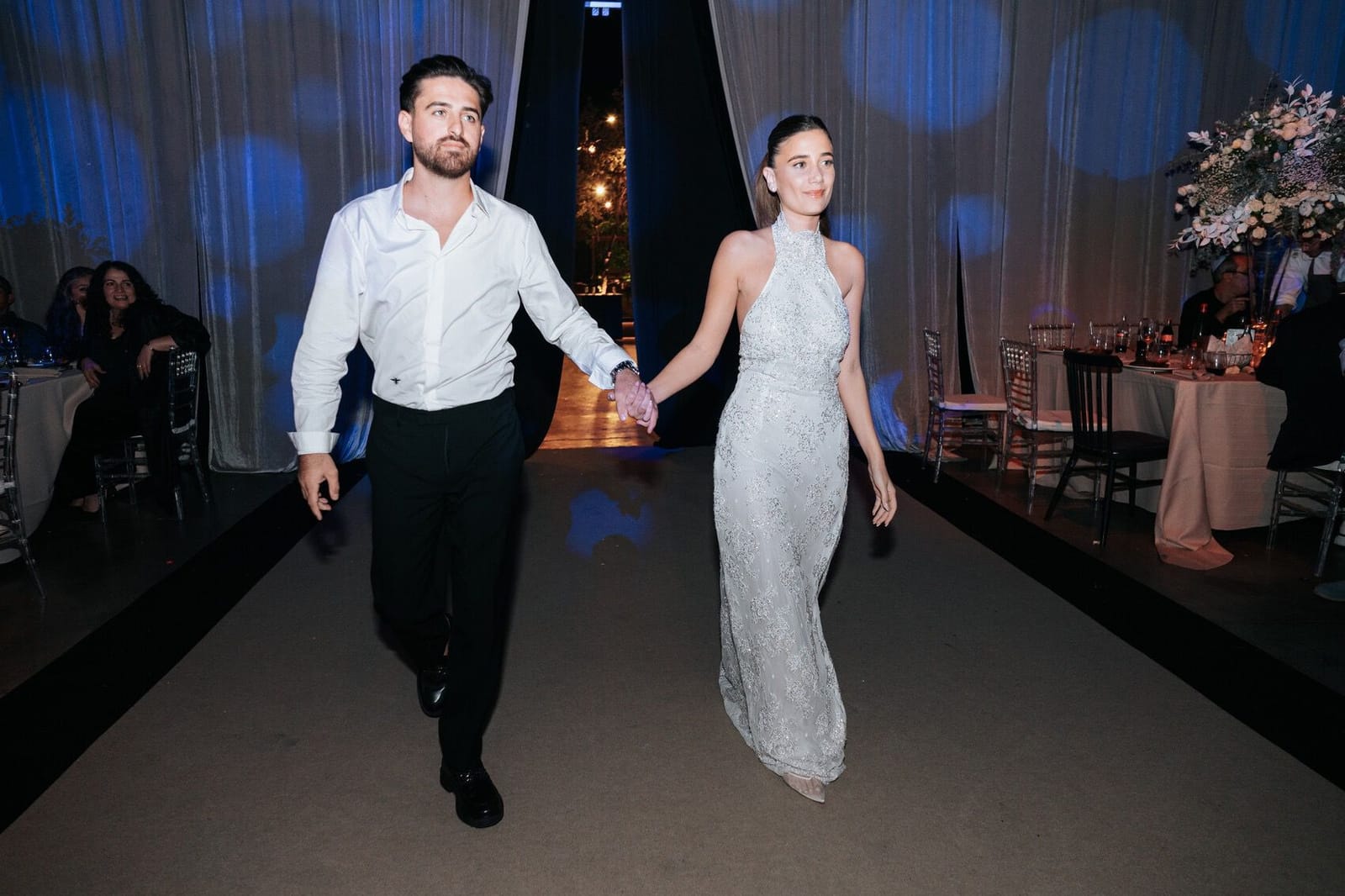 Un couple marchant main dans la main sur un podium sombre, lui en chemise blanche, elle en robe claire, lumières bleues derrière