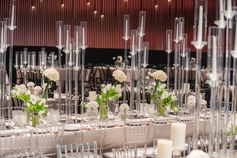 Many tall clear glass candle holders on a long table with white flowers and clear chairs before a reddish paneled wall
