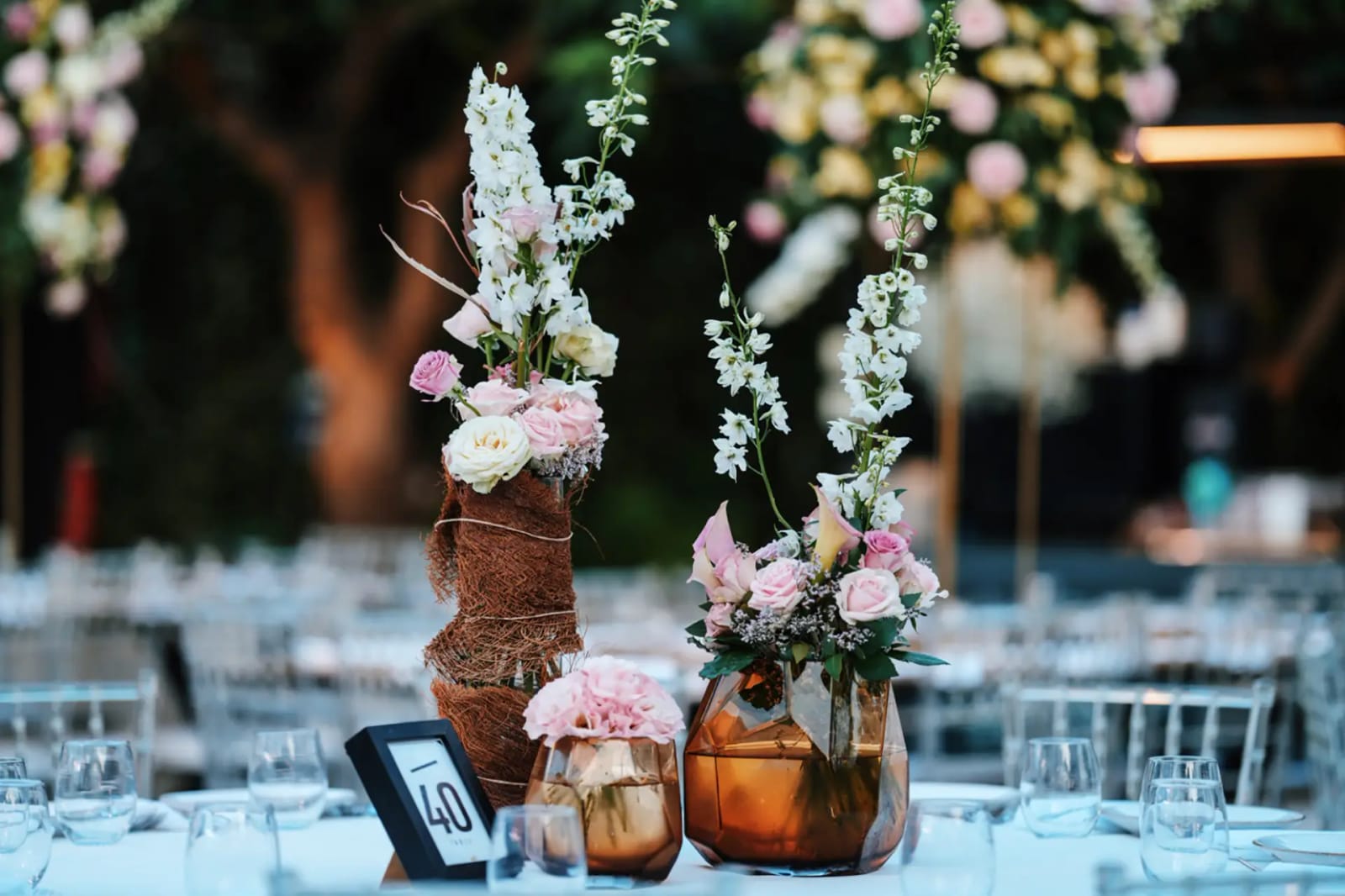 Copper and amber vases of pink roses and white delphinium beside a table number, with set tables blurred behind