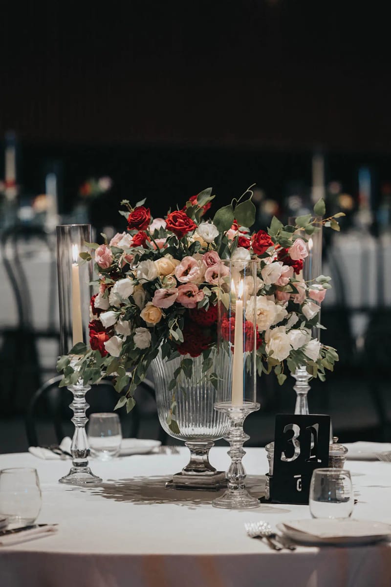Centre de table de roses rouges, roses et blanches dans un vase de verre sur pied, avec bougies et numéro de table