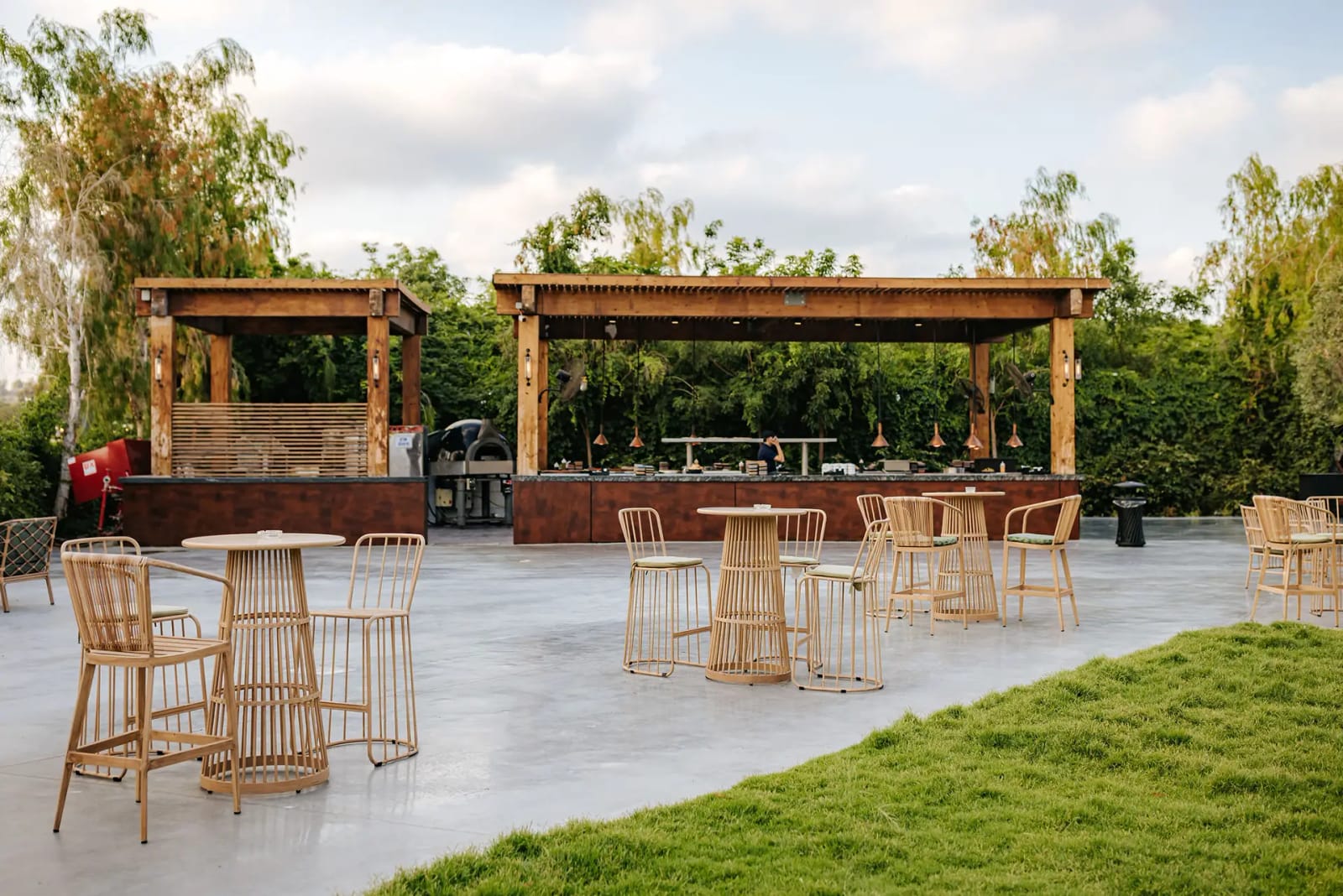 Deux pavillons de bar en bois sur une terrasse en béton avec tables hautes et tabourets en rotin, près d'une pelouse