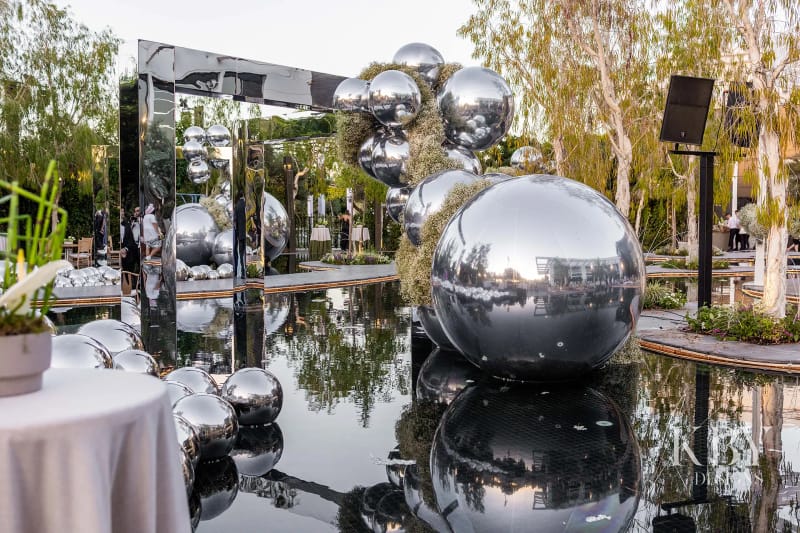 Silver mirror spheres of different sizes on dark reflective water, with a mirrored frame and trees behind