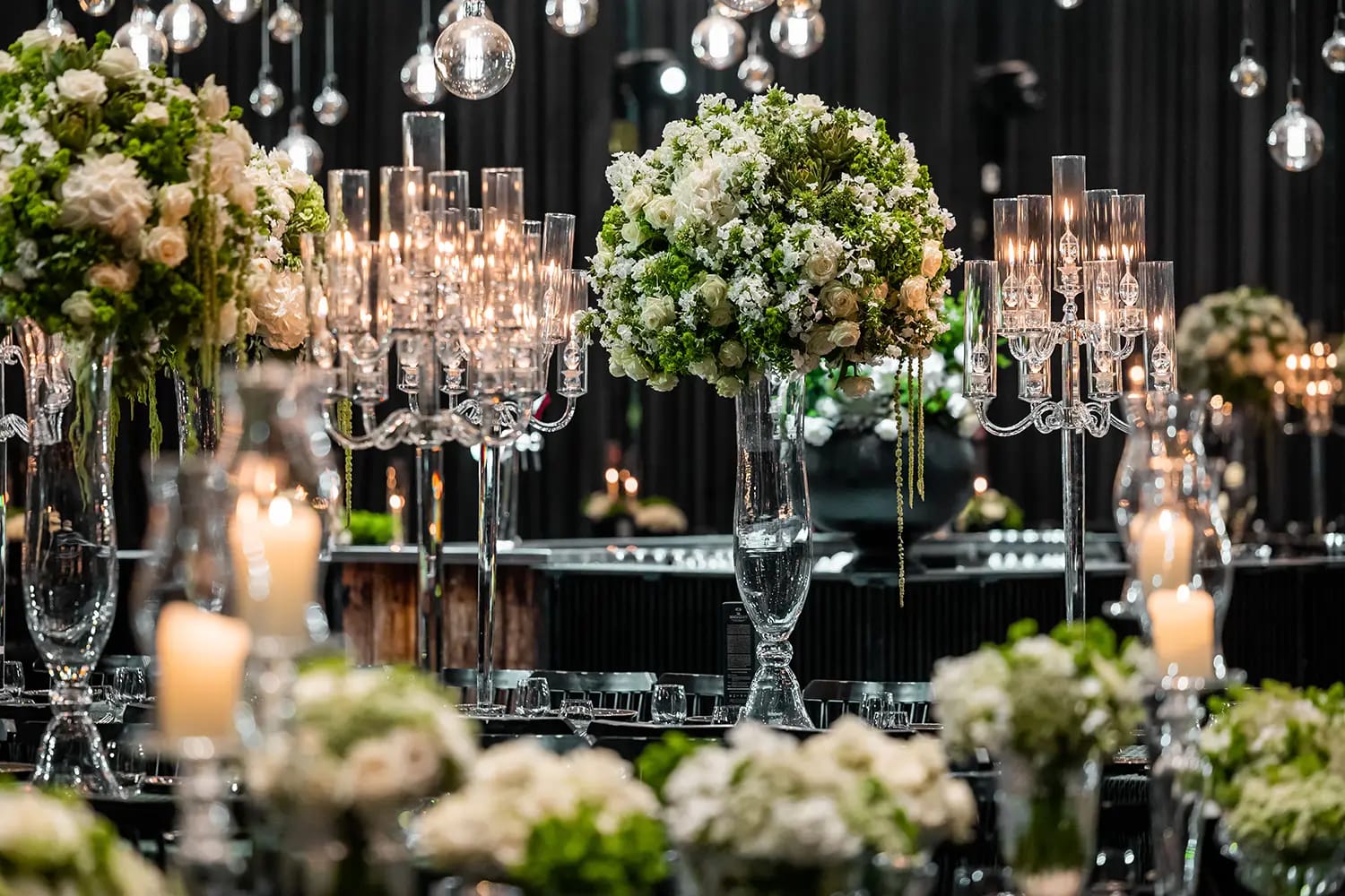 Candélabres en verre aux bougies allumées et fleurs blanches et vertes en grands vases, ampoules suspendues au-dessus
