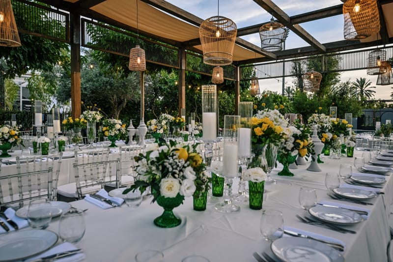 Long set table with green glass vases and yellow and white flowers under a pergola with hanging rattan lanterns