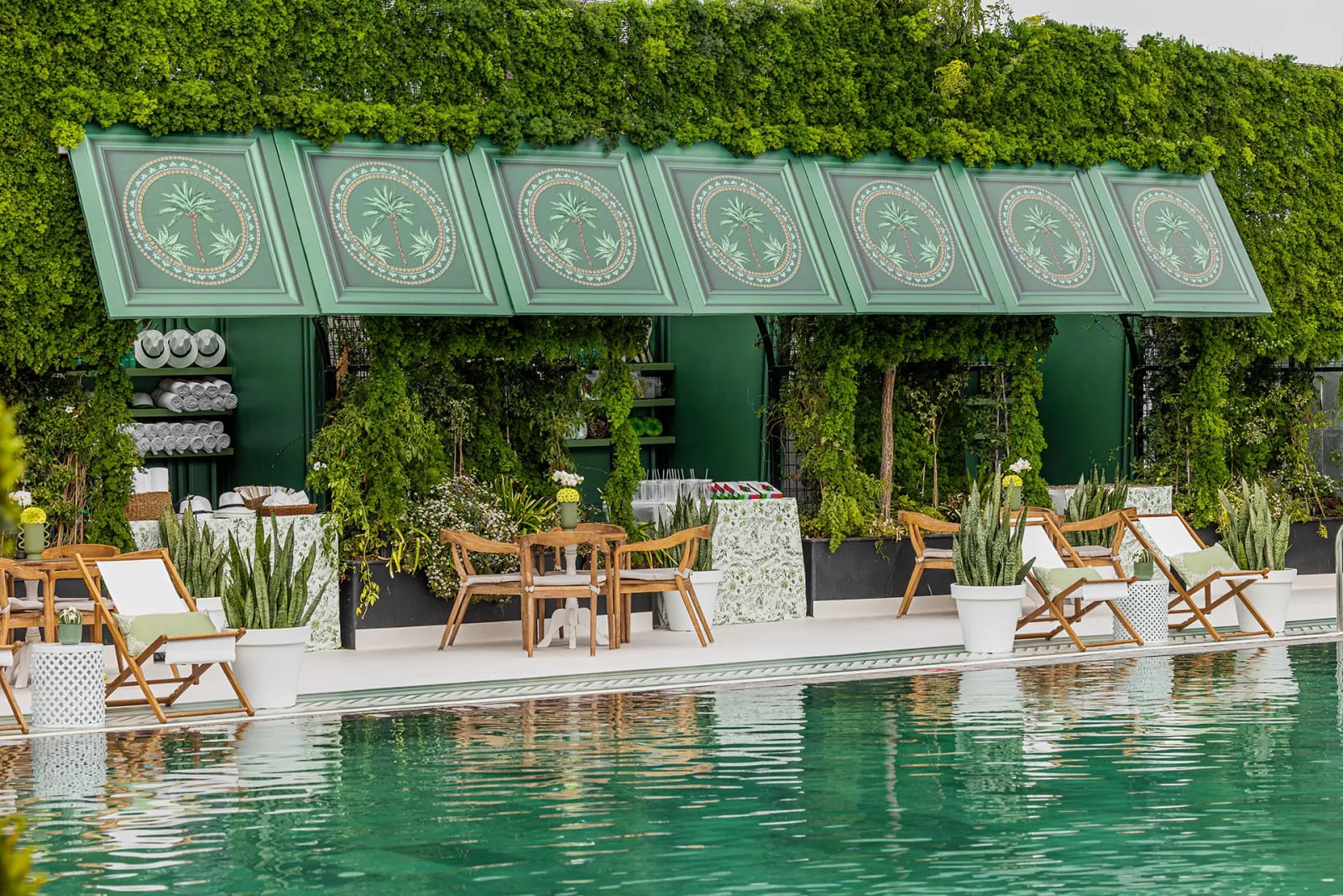 A poolside pavilion with ornate green panels, a hedge wall and white loungers beside a turquoise pool