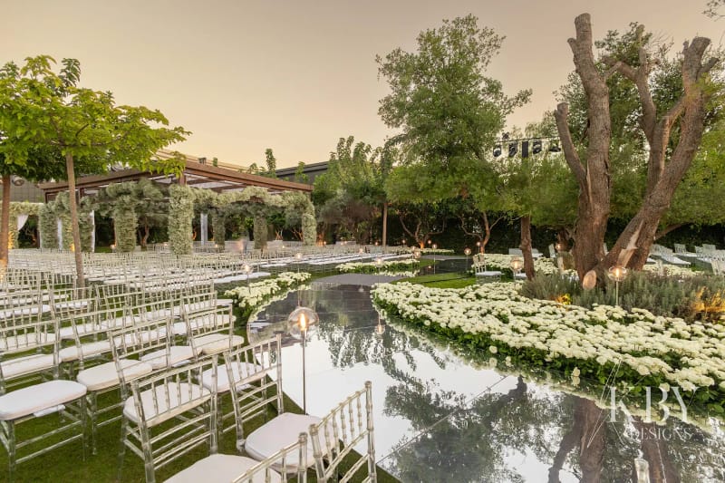 Clear chairs on both sides of a curving mirrored aisle edged with white flowers, among olive trees at dusk