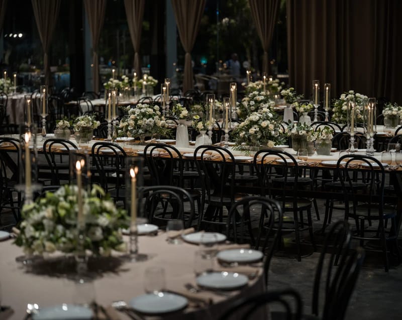 Round and long tables with green-and-white floral arrangements and candles, black chairs and brown curtains