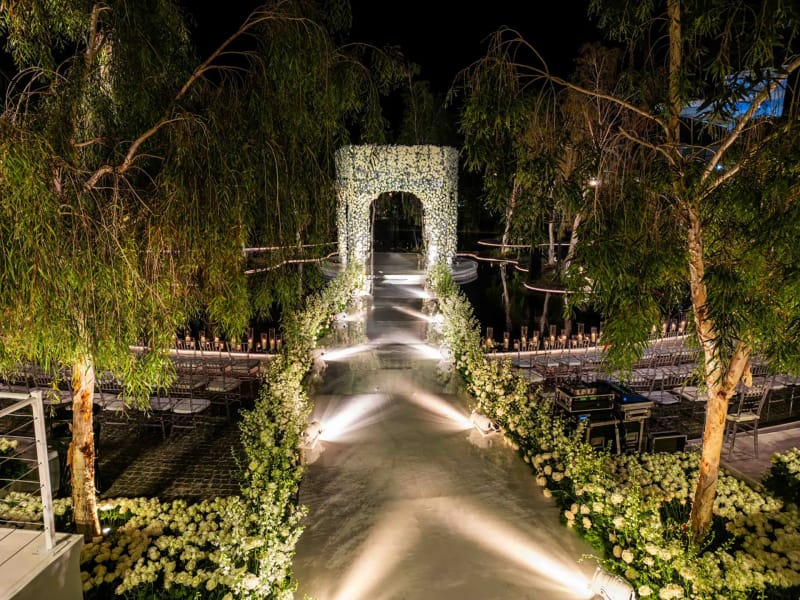 Arche de fleurs blanches au bout d'une allée éclairée entre des arbres, avec des bordures fleuries de chaque côté, de nuit