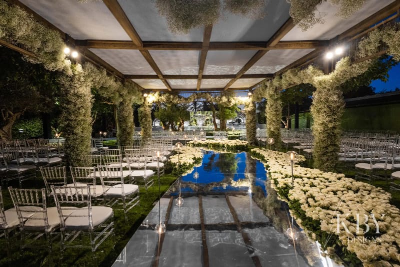 Wooden pergola with columns covered in white flowers over a mirrored aisle in the evening, with lights and clear chairs