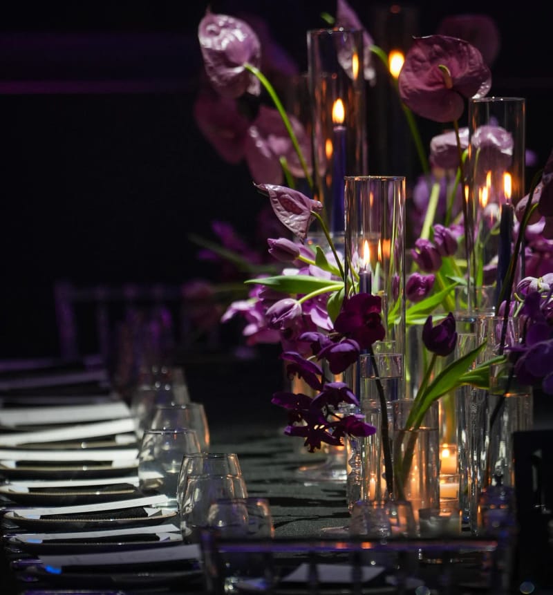 Purple anthurium and orchids in glass cylinders with candles, along a long dark table