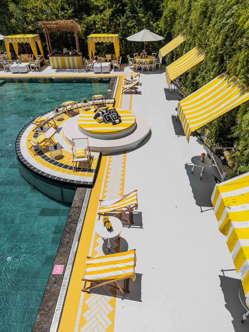 White walkway with yellow tile edging along the pool, striped awnings and a round seating platform by a green wall