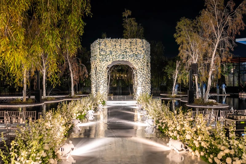 Arche de fleurs blanches au bout d'une allée éclairée entre des arbres, avec des rangées de chaises de chaque côté, de nuit