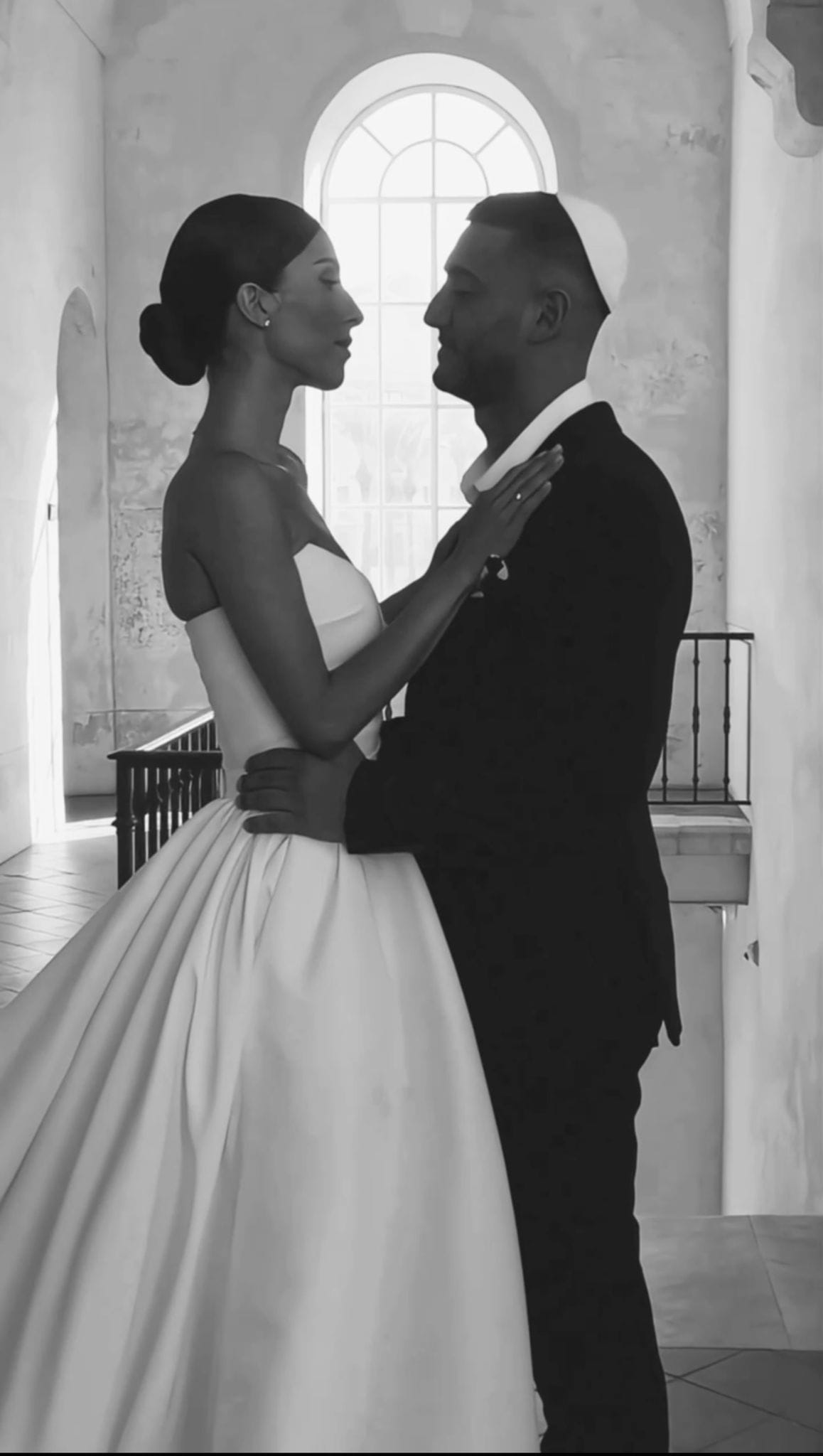 Image en noir et blanc d'un couple très proche devant une fenêtre en arc, elle en robe de bal, lui en costume sombre et kippa